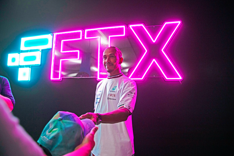 A man in a white Mercedes F1 team shirt stands in front of a large pink and cyan neon FTX sign on a dark background, receiving a teal cap from another person in the foreground.